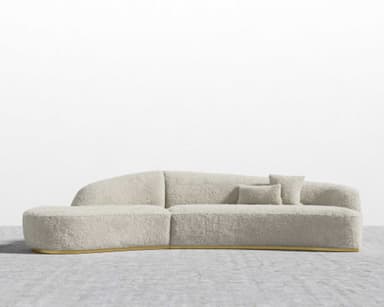 A contemporary, sculptural sofa featuring a deeply curved and organic silhouette, fully upholstered in a textured, cream-colored boucle fabric. The design includes integrated backrest elements that flow seamlessly from the main body. It is supported by a recessed, polished brass plinth base, giving it a floating appearance. — 3D model