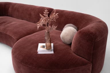 A contemporary curved sectional sofa upholstered in a rich, russet-toned velvet fabric. The sofa features a low-profile, modular design with gently rounded edges and continuous, integrated armrests and backrests. Its sinuous form is composed of at least two connected sections, creating an inviting and cohesive seating arrangement with no visible legs, resting directly on the floor. — 3D model