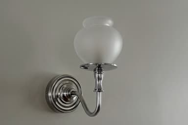 A polished nickel or chrome wall sconce featuring a frosted glass shade in an urn-like shape. The arm is gently curved, extending from a circular, fluted backplate, providing ambient or accent lighting. — 3D model