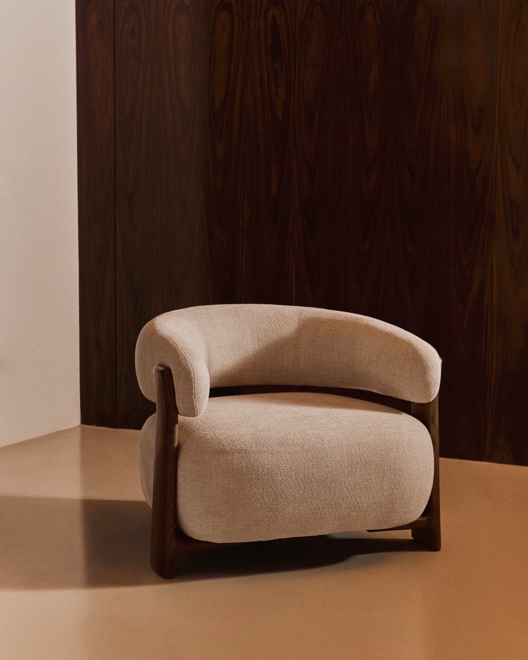 An upholstered armchair featuring a curved barrel-style backrest that seamlessly integrates into armrests, supported by a distinct exposed dark wood frame. The seat is thickly cushioned, and the entire piece is covered in a light beige, textured fabric. — 3D model