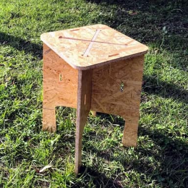 A small, four-legged stool constructed from oriented strand board (OSB). The top surface is square with rounded corners and features incised cross-shaped lines. The legs are trapezoidal in shape with cutout sections and a circular hole at the bottom of each, possibly for assembly or decoration. The stool has a minimalist, modern aesthetic and appears to be designed for flat-pack assembly. — 3D model