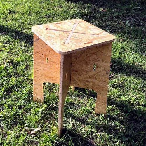 A small, four-legged stool constructed from oriented strand board (OSB). The top surface is square with rounded corners and features incised cross-shaped lines. The legs are trapezoidal in shape with cutout sections and a circular hole at the bottom of each, possibly for assembly or decoration. The stool has a minimalist, modern aesthetic and appears to be designed for flat-pack assembly. — 3D model