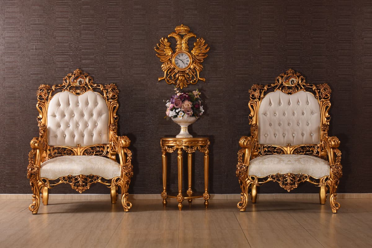 A pair of highly ornate, gilded armchairs with tufted cream upholstery, embellished with decorative carvings and crystal buttons, flanking a matching gilded accent table topped with a vase of flowers and situated against a textured dark brown wall, beneath an ornate gilded wall clock. — 3D model
