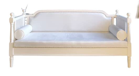 A white daybed featuring a solid wood frame, cane webbing details on the arms, and an upholstered back panel with nailhead trim. It includes a single long cushion and two cylindrical bolster pillows. — 3D model