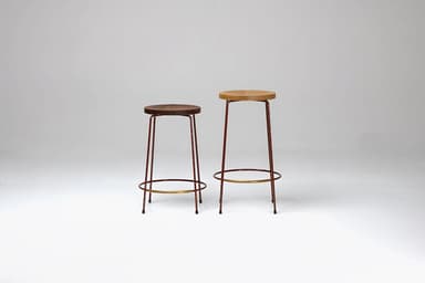 A minimalist bar stool featuring a round, natural oak wood seat and a slender four-legged metal base. The legs are a dark reddish-brown hue, tapering slightly towards the floor, and are reinforced with a circular brass-toned metal footrest. — 3D model