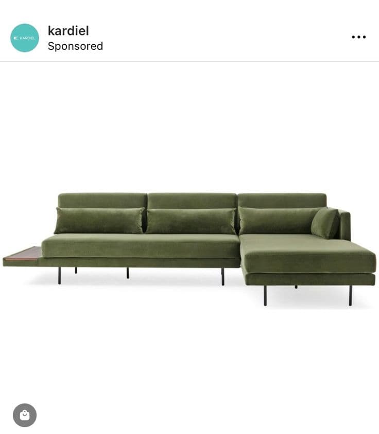 A modern sectional sofa upholstered in olive green velvet, featuring an L-shaped configuration with a right-hand chaise lounge extension. The design incorporates a low profile, slim black metal legs, and a rectangular wooden side table seamlessly integrated at one end. It is furnished with loose back cushions and small bolster pillows. — 3D model