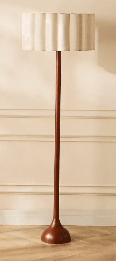 A freestanding floor lamp featuring a tall, slender wooden stem and a matching round, turned wooden base. The lamp is topped with a cylindrical drum shade crafted from a light-colored fabric, distinctively shaped with a scalloped, wavy rim at both the top and bottom edges. The fabric shade appears to be beige or cream, while the wood components have a warm, reddish-brown finish. — 3D model