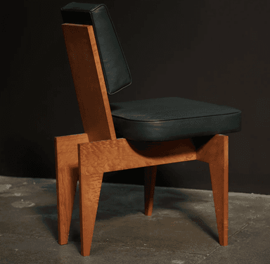 An armless chair featuring a distinctive geometric wooden frame with a light-colored, possibly burled, wood grain. The seat and separate backrest are upholstered in a dark green material, likely leather or a high-quality synthetic, offering a plush appearance. The wooden base is composed of thick, angular planks forming the legs and supporting structure, with a splayed design that tapers slightly towards the floor. The chair has a low profile backrest, suggesting an accent or side chair function. — 3D model