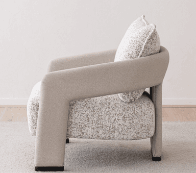 A contemporary upholstered armchair featuring a rounded silhouette and a neutral-toned, speckled fabric. The chair includes integrated, curved armrests and an upholstered backrest, complemented by a separate, plush back cushion with piped edges. The seat appears integrated, and the chair stands on short, upholstered legs or a low, concealed base, suitable for modern interior settings. — 3D model