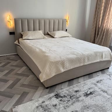 A modern upholstered platform bed featuring a tall headboard with vertical channel tufting. The entire bed frame, including the headboard and side rails, is covered in a neutral-toned fabric, likely beige or light gray. The bed is presented with a white mattress, two matching pillows, and a textured white duvet/quilt. It has a low-profile design, sitting on small, recessed feet. — 3D model