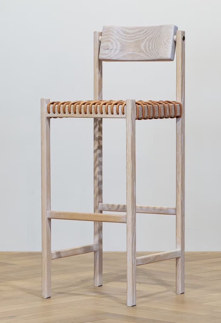 A tall bar stool with a light wood frame and a woven seat and backrest. The frame consists of four vertical legs connected by horizontal stretchers. The seat is made of intertwined leather straps, and the backrest is a solid, slightly curved wooden piece attached to the upper part of the frame. — 3D model