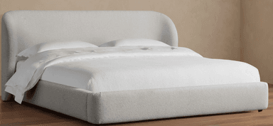 A close-up view of a modern upholstered bed frame, featuring a softly curved, enveloping headboard and side rails covered in a textured, light-colored boucle fabric. A white pillow and bedsheet are visible, indicating its function. — 3D model
