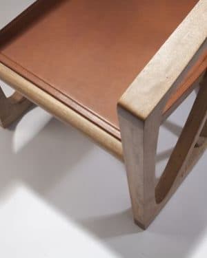 A contemporary armchair crafted from light-colored solid wood, featuring a gracefully curved backrest and integrated armrests. The chair incorporates a geometric, sculptural base with multiple intersecting wooden struts providing support. A contrasting reddish-brown leather cushion forms the seat, complementing the natural wood tone. — 3D model