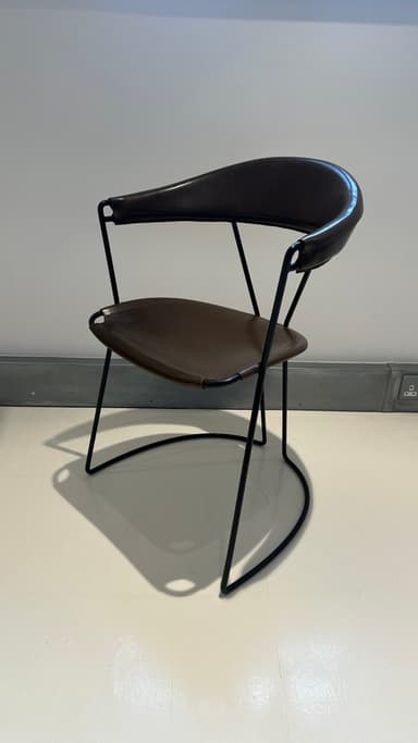 A contemporary armchair with a dark brown leather seat and backrest supported by a slender black metal frame. The frame features a curved base that extends into the legs, providing stability and a dynamic visual element. The seat and backrest are subtly contoured for comfort and appear to be constructed from stitched leather panels. — 3D model