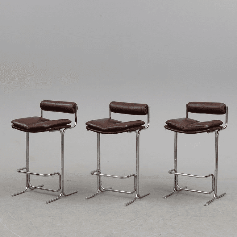 A modern bar stool featuring a gleaming tubular chrome frame and upholstered in rich brown leather. It has a padded square seat and a small, padded backrest for comfort, along with an integrated footrest. — 3D model