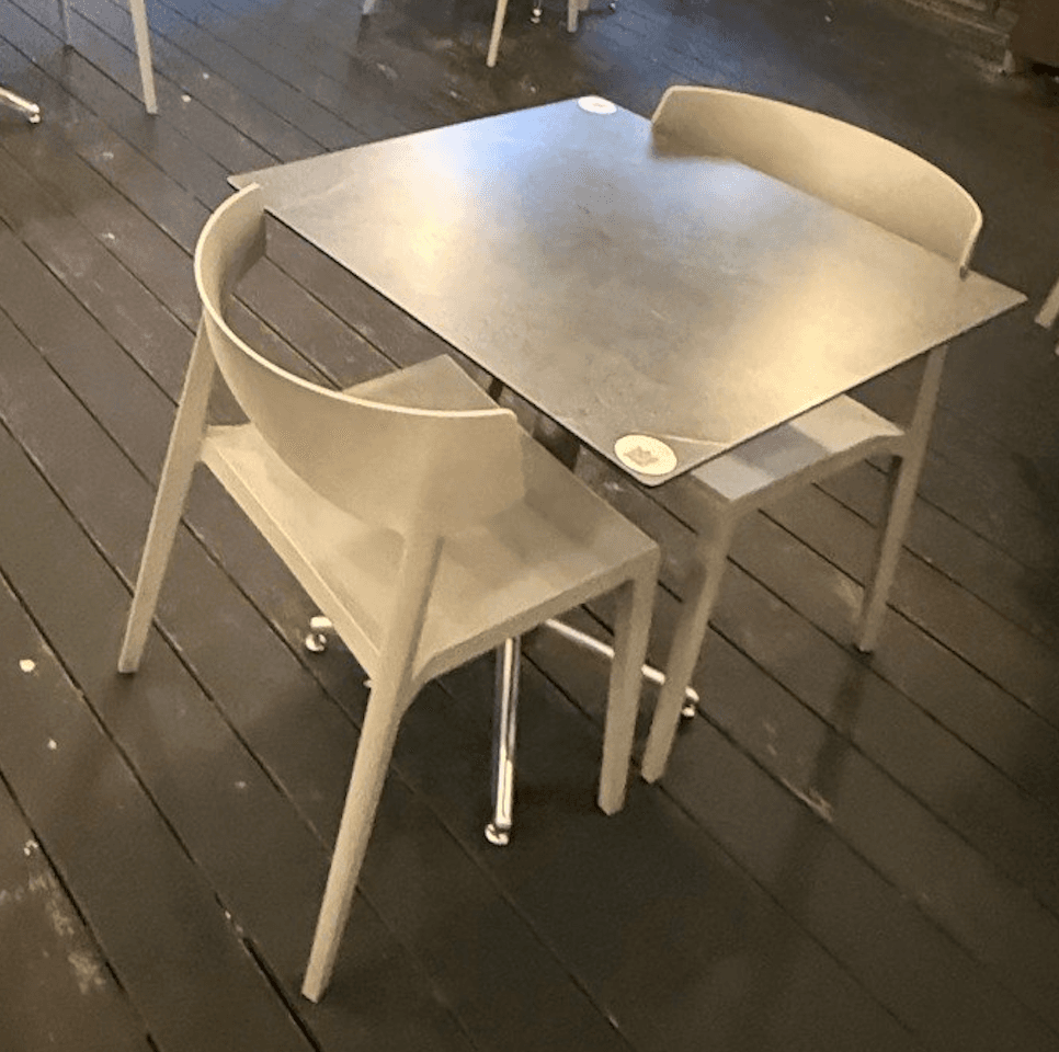 A modern dining set comprising a square tabletop and two chairs. The tabletop has a dark, matte finish and is supported by a central pedestal base with four feet. The chairs are molded, with a simple, curved backrest and four legs. The overall design suggests a contemporary aesthetic suitable for compact dining areas or cafes. — 3D model