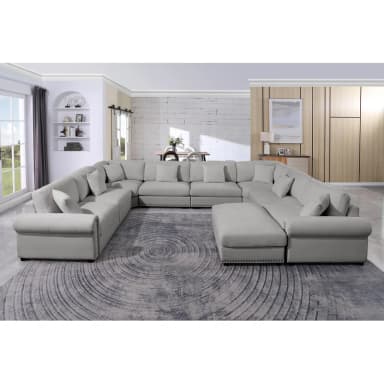 A large, light gray sectional sofa with a U-shaped configuration and an attached ottoman. The sofa features multiple seating modules, plush cushions, and decorative nailhead trim along the base. It includes several throw pillows for added comfort and style. — 3D model