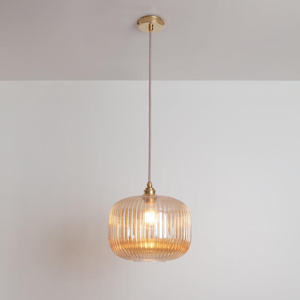 A single amber ribbed glass pendant light featuring brass hardware for mounting and suspension. The glass shade is a rounded rectangular form with vertical fluting, enclosing a visible light bulb. A fabric-covered electrical cord connects the shade to a circular brass canopy, providing a sleek and modern design with warm, diffused illumination. — 3D model