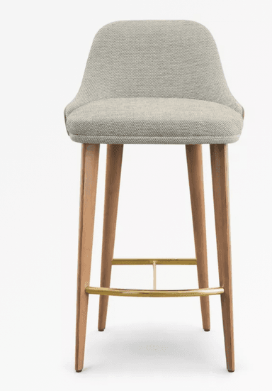 A contemporary bar stool featuring a light grey upholstered seat and low back, supported by four tapered wooden legs with polished brass footrests. — 3D model