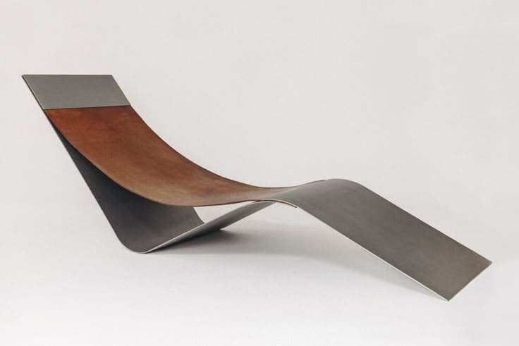 A modern chaise lounge with a minimalist design, featuring a curved frame that supports a distinct seating surface. The frame appears to be made of metal, possibly brushed or polished, and transitions into a material that resembles leather or a similar tanned hide for the seating area. The overall form is sleek and organic, designed for relaxation. — 3D model