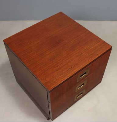 A compact, three-drawer wooden nightstand or side table, characterized by its reddish-brown finish and distinctive brass campaign-style recessed pull handles, supported by a recessed plinth base. — 3D model