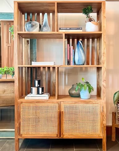 A tall, rectangular bookshelf crafted from light-toned natural wood, featuring multiple open shelving compartments in the upper section and two cabinet doors with woven cane webbing in the lower section. The upper section includes slatted vertical elements along the sides and some internal dividers, creating an airy display space. The lower cabinets have simple, rectangular fronts with the cane infill, resting on a recessed plinth-like base or integrated short legs. — 3D model