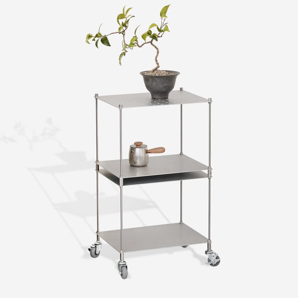 A three-tiered metal cart with casters. The top shelf features a perforated pattern. The cart is made of thin metal rods and shelves, giving it a minimalist and industrial aesthetic. It is shown with a bonsai plant on the top shelf and a metal pot with a wooden handle on the middle shelf. — 3D model