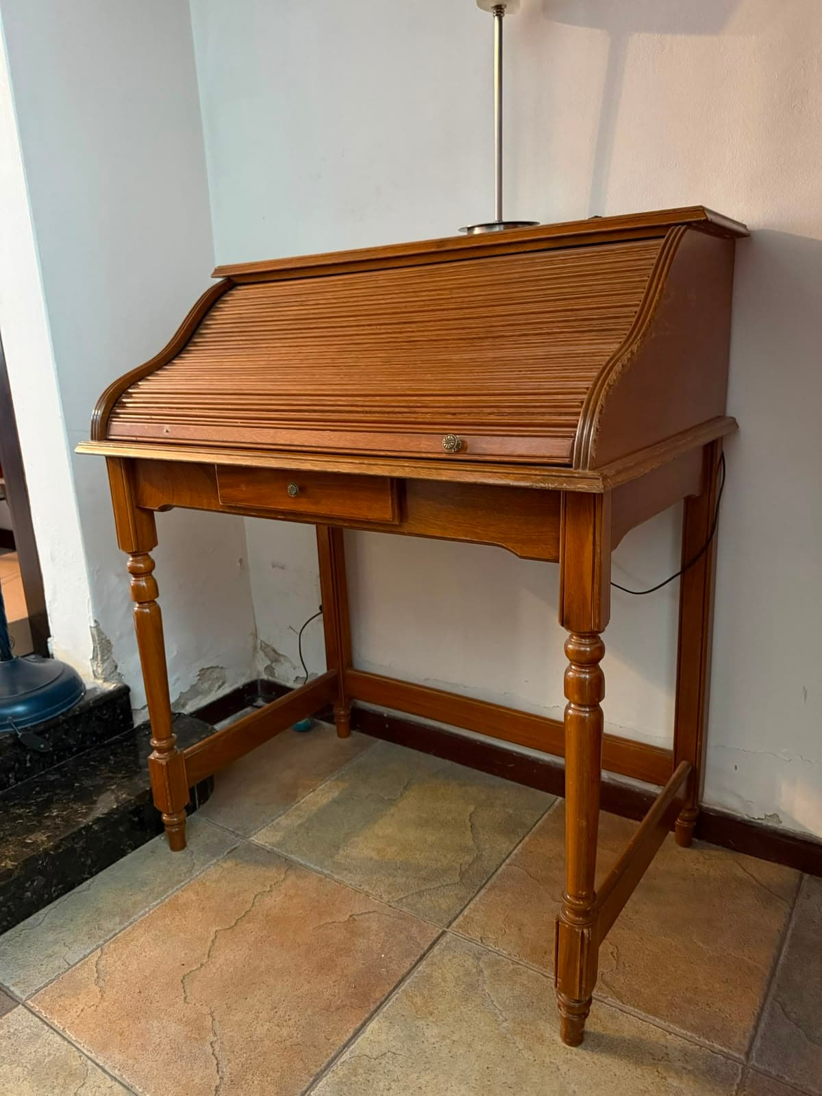 A traditional wooden roll-top secretary desk featuring a slatted roll-top mechanism that conceals the work surface. The desk includes a single central drawer below the roll-top section, equipped with a small wooden knob. It stands on four turned legs, connected by stretchers on the sides and front, suggesting a classic or antique aesthetic. The desk is finished in a medium brown wood tone. — 3D model