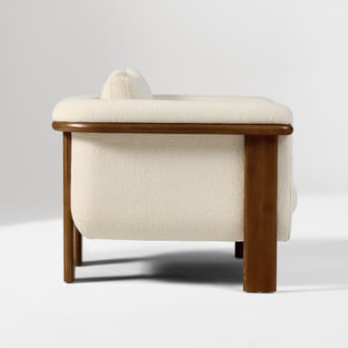 A contemporary armchair featuring a solid wood frame with natural wood grain visible. The seating and backrest are upholstered in a textured, light-colored fabric. The design incorporates a rounded aesthetic with distinct block-style wooden legs and integrated armrests that extend from the frame. — 3D model