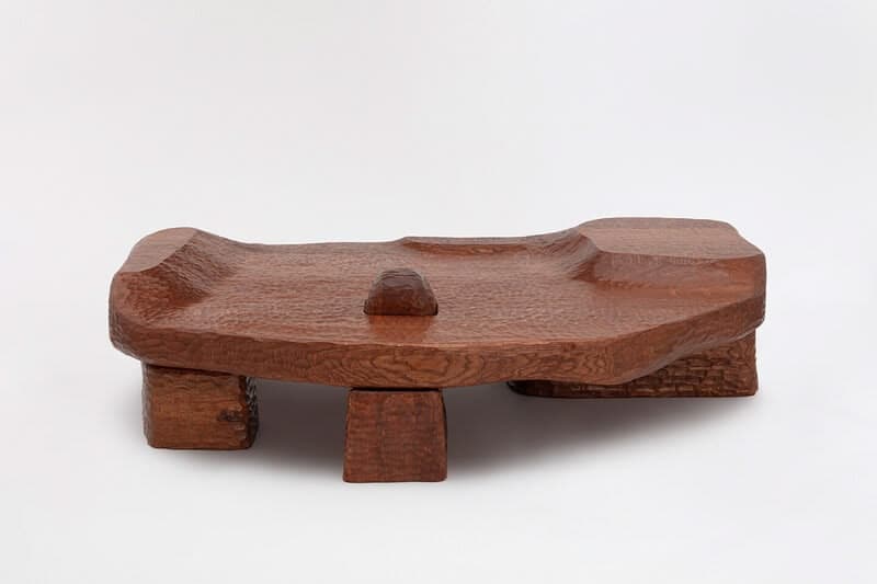 A unique, low-profile coffee table carved from wood. The tabletop features an organic, undulating surface with a textured finish, resembling natural erosion or flowing water. It is supported by four block-like legs, also made of wood. A single, small rectangular block rises from the center of the tabletop. The overall aesthetic is rustic and sculptural, with visible tool marks and a rich wood grain. — 3D model