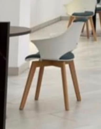 A contemporary armchair featuring a white molded plastic shell with integrated armrests and a cut-out back. The seat is fitted with a dark grey upholstered cushion. It stands on four splayed and tapered light-toned wooden legs. — 3D model