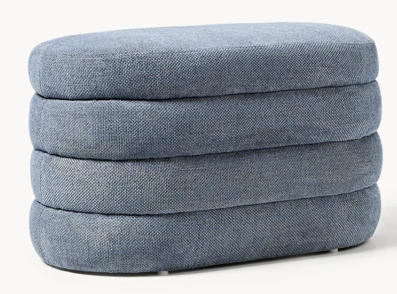 A contemporary ottoman with a rounded, elongated rectangular form. It is constructed from multiple stacked, cushion-like segments, each upholstered in a textured blue fabric. The segments create a layered appearance, and the overall shape is soft and organic. It appears to have a low-profile base. — 3D model