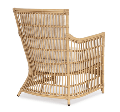 A light brown natural rattan lounge armchair featuring a curved, open-weave frame. It is fitted with thick, removable, light beige upholstered seat and back cushions. The chair has a rounded back and arms, with vertical rattan slats forming the back and side panels. The legs are integrated into the curved frame, with the rear legs slightly flared. The overall aesthetic is natural and relaxed. — 3D model