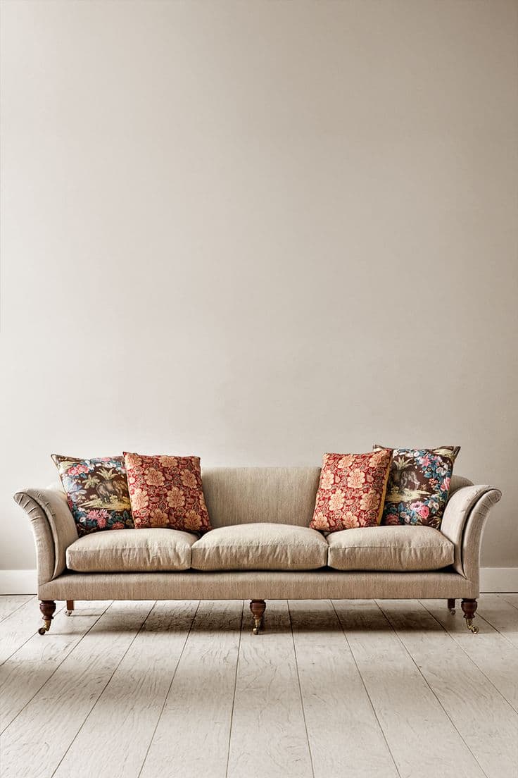 A traditional three-seater sofa upholstered in a neutral beige fabric, featuring classic rolled arms and dark turned wooden legs with brass casters. The sofa includes three separate seat cushions and several loose, patterned throw pillows. — 3D model