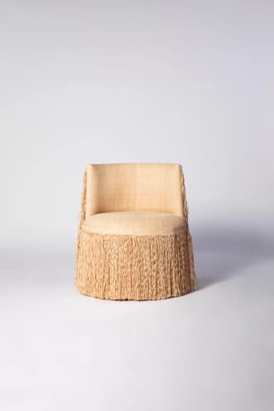 An armless accent chair featuring a distinctive design with a fully fringed base and lower back, upholstered in a light beige textured fabric. The chair has a round seat and a low, rectangular backrest, with the long, natural-toned fringe extending from beneath the seat and down the sides of the backrest, giving it a playful and contemporary appearance. — 3D model