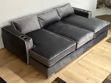 A modern sectional sofa with a chaise lounge extension on one side. The sofa features multiple seat cushions and back cushions, upholstered in a soft, plush gray fabric. It has a piped trim in a lighter contrasting color along the edges of the cushions and frame. Two integrated cup holders are visible on the armrests. The base appears to be a solid structure, possibly with decorative metal feet. — 3D model