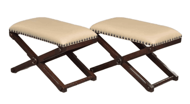 A campaign-style X-frame ottoman featuring a dark wood base and an upholstered seat. The seat is covered in a light, cream-colored material and secured with decorative nailhead trim along the bottom edge. The X-shaped wooden legs are connected by turned stretchers, and the overall design suggests a traditional or vintage aesthetic. — 3D model