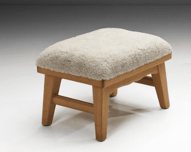 A rectangular stool featuring a thick, plush, cream-colored sheepskin upholstery top over a natural light-toned wooden frame. The frame has visible grain, sturdy splayed legs, and a subtly scalloped apron detail on the long sides. — 3D model