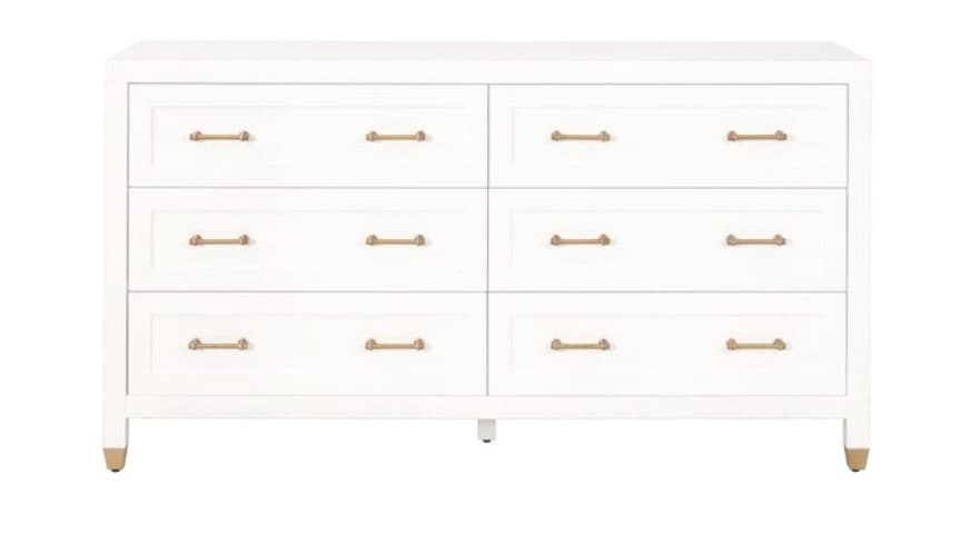 A white dresser with six drawers, arranged in two columns of three drawers each. The dresser features horizontal brass-colored handles on each drawer. The top surface is flat and the sides have a slightly rounded profile. It stands on four legs, with the front legs having brass-colored accents at the base. — 3D model