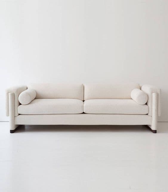A modern, two-seater sofa with a clean, minimalist design. It features a rectangular frame, upholstered in a light-colored, textured fabric. The sofa has a low profile, with distinct square armrests and a single bench seat cushion. Two cylindrical bolster pillows are placed at either end of the sofa. The legs are short and dark-colored, providing a subtle contrast to the upholstery. — 3D model
