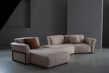A contemporary modular sectional sofa upholstered in a light beige textured fabric, featuring a left-hand chaise lounge and a right-hand armrest with a distinctive dark framed detail. It is adorned with several brown and beige throw pillows. — 3D model