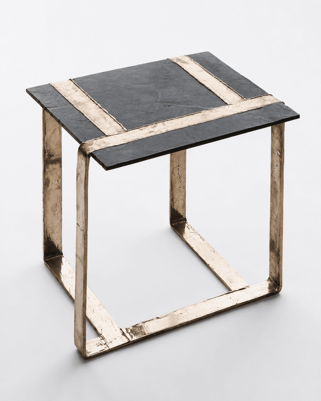 A square side table featuring a dark, textured slate top and a robust, open-cube base constructed from raw, cast bronze. The slate tabletop is inset with two perpendicular strips of the same bronze material, creating a distinctive cross pattern. The bronze frame exhibits a deliberately unfinished, organic texture, emphasizing its handcrafted nature and solid construction. The overall aesthetic is heavy, raw, and industrial, typical of Brutalist design influences. — 3D model