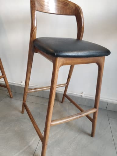 A contemporary bar stool featuring a warm-toned wooden frame, a curved backrest, and a padded black upholstered seat. — 3D model
