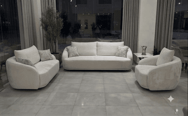 A modern, light beige three-seater sofa with a distinctive curved silhouette, upholstered in a subtly textured fabric, featuring three seat cushions, two back cushions, and two patterned throw pillows. — 3D model