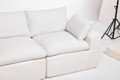 A modular sectional sofa with an attached ottoman. The sofa features deep, plush cushions and a low profile. It is upholstered in a textured white fabric. The configuration shown includes a corner section, two seat cushions, and an ottoman. The design is contemporary and emphasizes comfort. — 3D model
