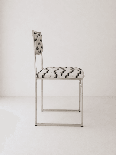 A contemporary armless chair featuring a minimalist, rectangular frame constructed from polished tubular metal. The seat and backrest are upholstered in a fabric with an abstract geometric pattern in shades of cream, black, and grey. The frame forms continuous loops for the legs and supports the seat and back. — 3D model