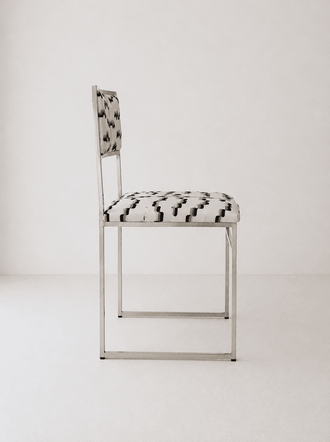 A contemporary armless chair featuring a minimalist, rectangular frame constructed from polished tubular metal. The seat and backrest are upholstered in a fabric with an abstract geometric pattern in shades of cream, black, and grey. The frame forms continuous loops for the legs and supports the seat and back. — 3D model
