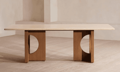 A long, rectangular dining table featuring a light-colored travertine top with rounded corners. The top is supported by two substantial, medium-toned wooden pedestal bases. Each base is characterized by vertical fluting or reeding and a large, semicircular cutout on its inner side, creating a sculptural and architectural aesthetic. — 3D model