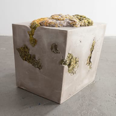 A cubic concrete ottoman with an irregular, textured top surface composed of braided and knotted yarn in shades of yellow, green, and grey. The concrete body features several irregular voids and openings, some of which reveal the same yarn material embedded within. — 3D model