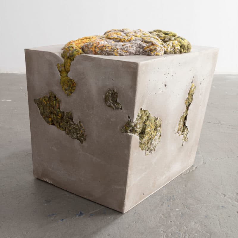 A cubic concrete ottoman with an irregular, textured top surface composed of braided and knotted yarn in shades of yellow, green, and grey. The concrete body features several irregular voids and openings, some of which reveal the same yarn material embedded within. — 3D model
